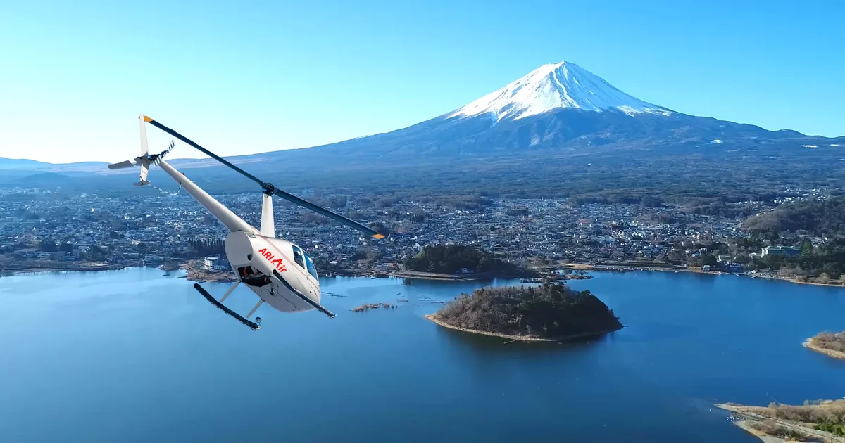 A small white helicopter flying over a lake with a wooded island, below snow-capped Mount Fuji on a clear day
