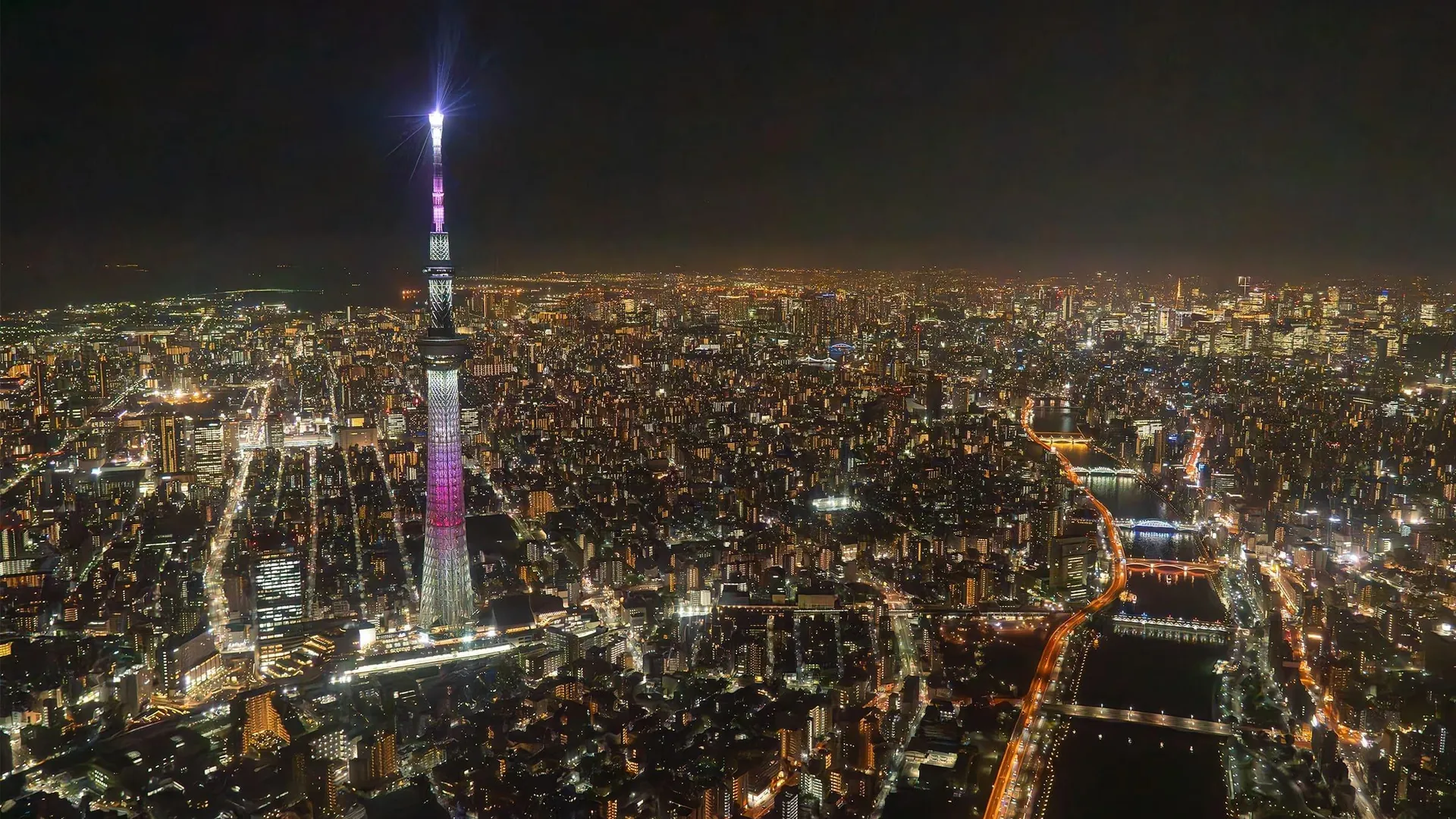 Tokyo Skytree lit up above the city lights and the Sumida River at night, seen from a helicopter
