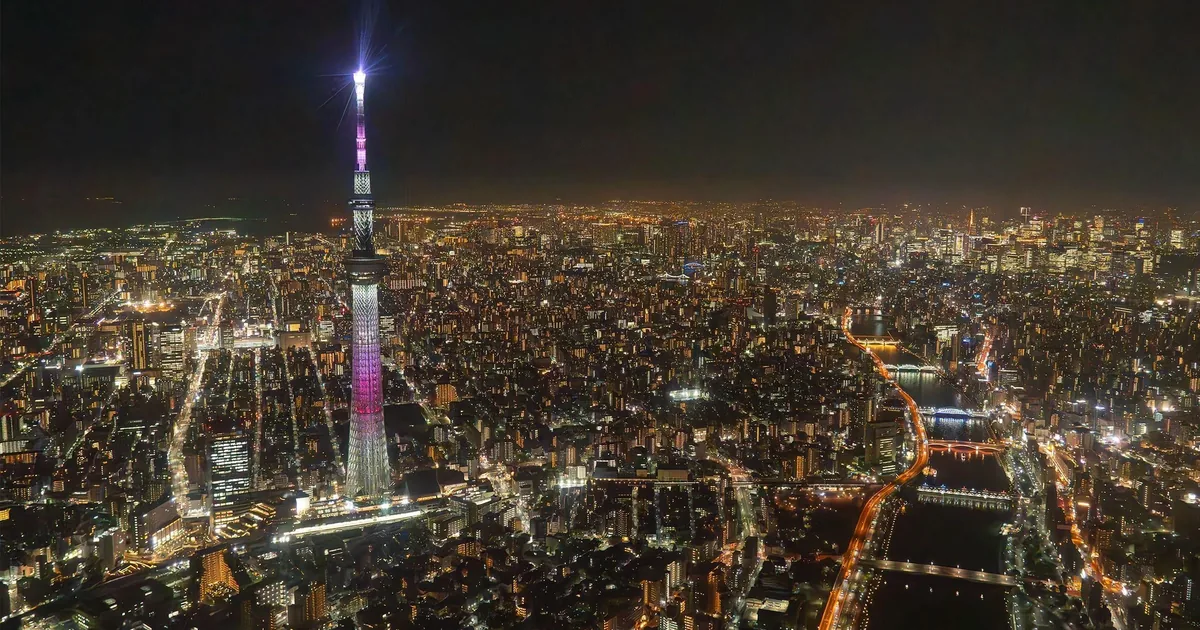 Tokyo Skytree lit up above the city lights and the Sumida River at night, seen from a helicopter